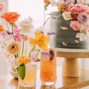 Pièce montée design décorée de fleurs sur un présentoir en bois, cocktails dans des verres, et bouquet de fleurs jaillissant dans les tons pastel