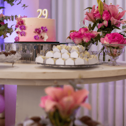 Table d'anniversaire où se trouvent un gâteau rose décoré de fleurs violettes, des bouquets de fleurs roses dans des coupes en verre, et des plateaux de petits fours